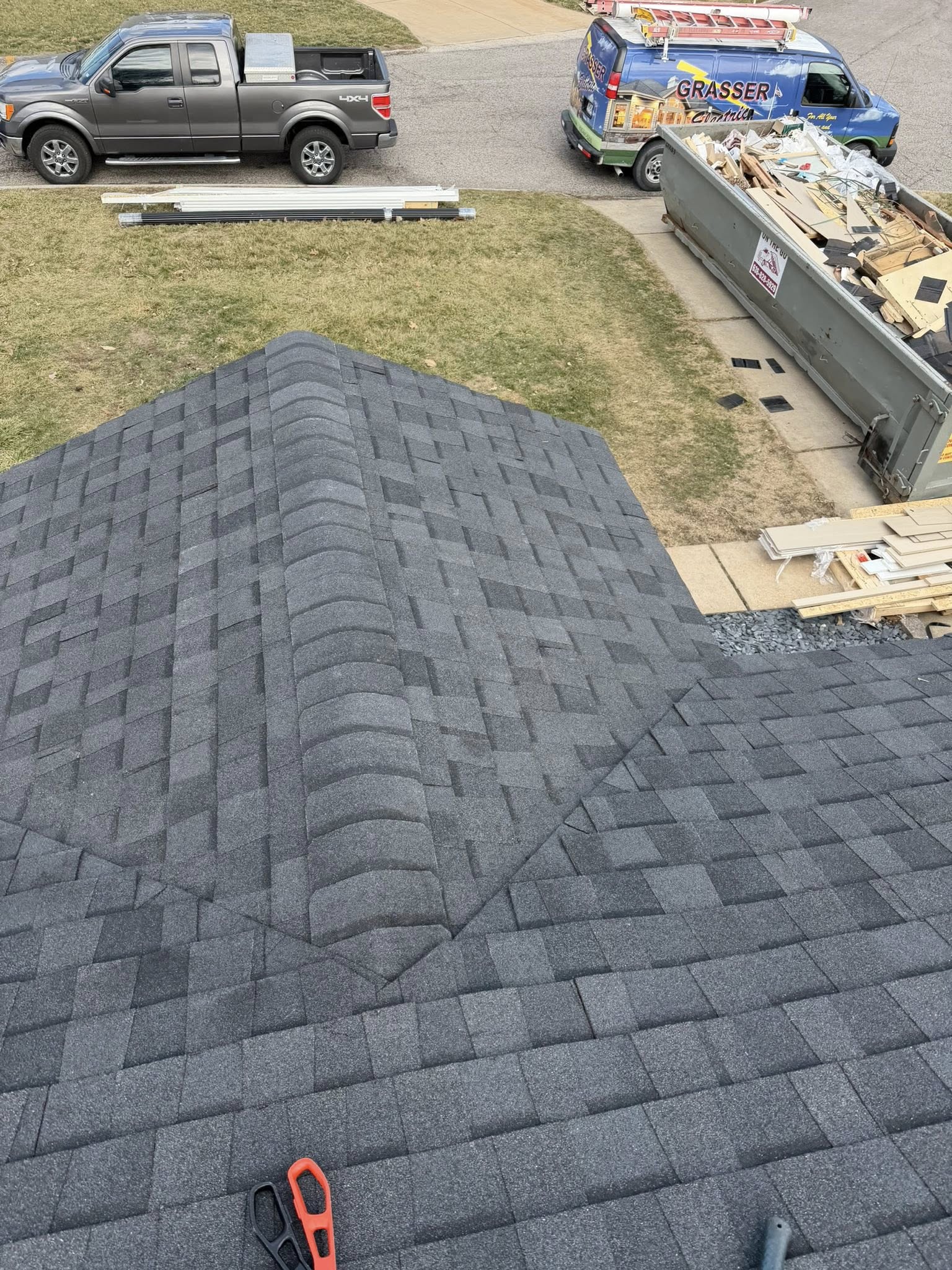 Aerial view of asphalt shingle roof installation in progress with work vehicles and materials on residential driveway